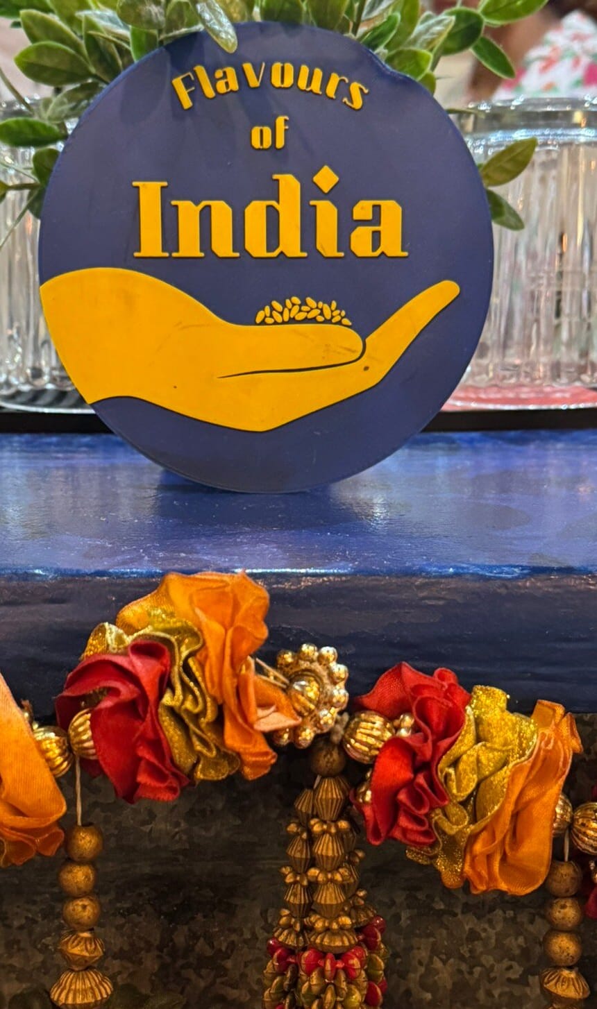 O emblema do Flavours of India em pé na prateleira azul do balcão, entre frascos e um festão de flores