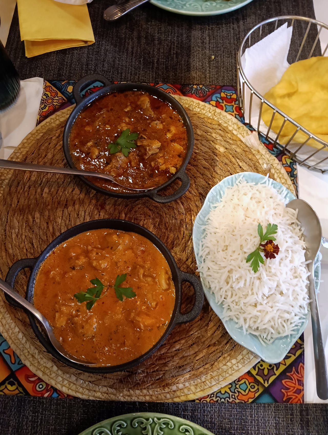 Dois caris em panelas de ferro, arroz basmati num prato verde em forma de folha e um cesto de papadums